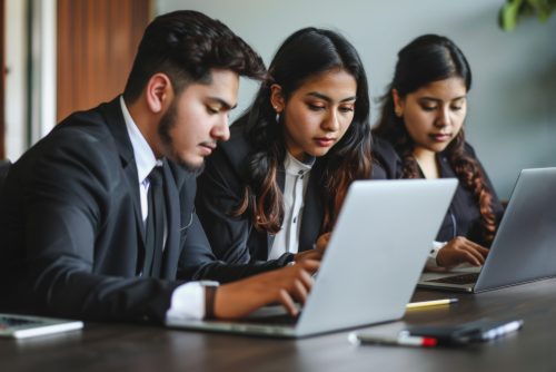 Three young Hispanic businesspeople are working on their laptops in a modern office setting. They are focused on their tasks, with one man in the foreground looking intently at his laptop while the two women beside him are also engaged in their work. The image captures a collaborative and professional atmosphere, highlighting the diversity and dynamism of the modern workforce.